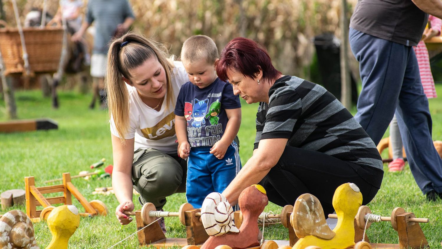 Two women and a child are outdoors, playing with wooden toys shaped like ducks and snails. The child is in the center, interacting with the toys. People and tall grass are visible in the background, suggesting a park or garden setting.