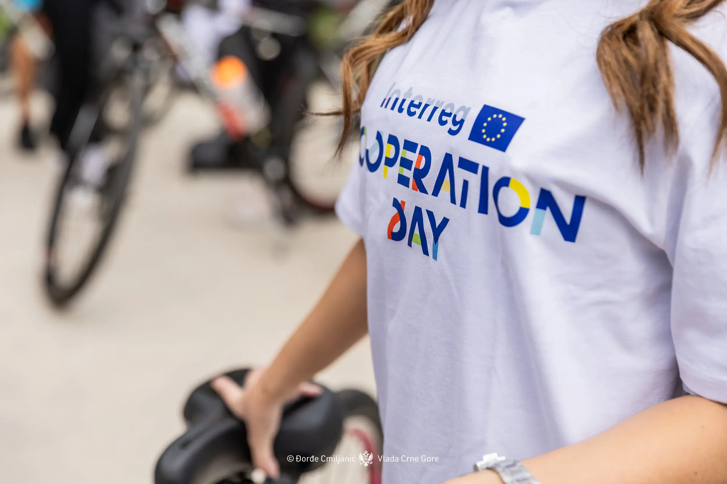 Person wearing a white T-shirt with "Interreg OPERATION DAY" in colorful letters. They are holding a bicycle seat, and blurred bicycles are visible in the background.