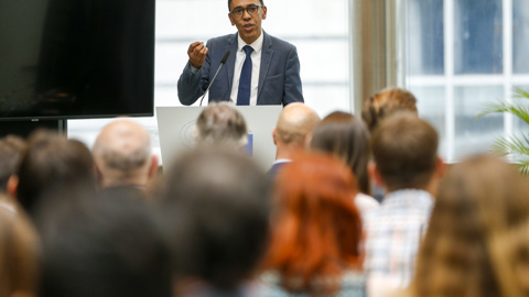 A man in a suit and tie speaks at a podium in front of an audience in a bright room with large windows. The audience is seated and listening attentively.