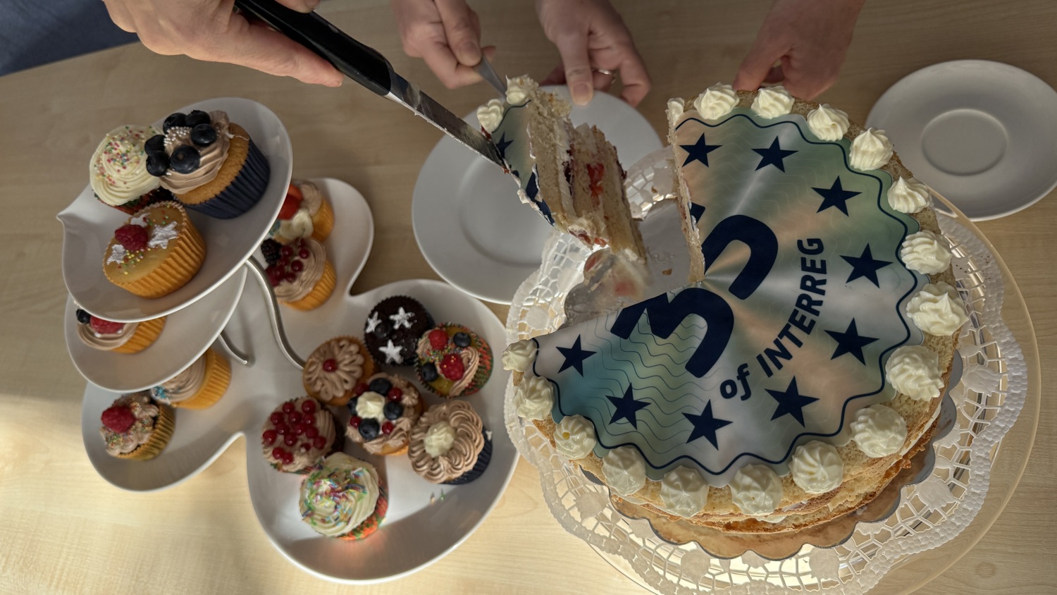 A person slices a cake decorated with a “35 of INTERREG” design while another person holds a plate; assorted decorated cupcakes are displayed nearby on a tiered tray.