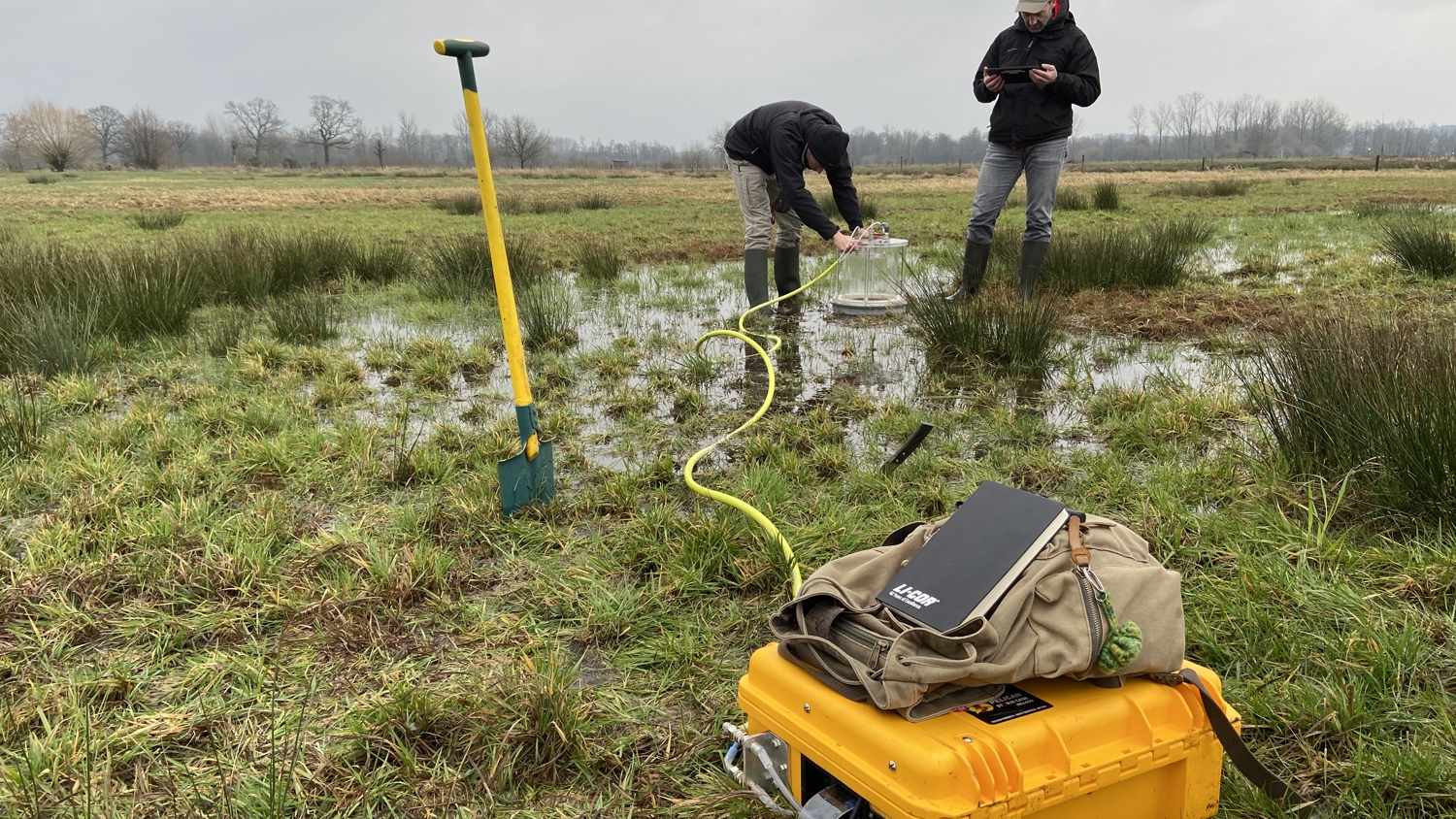 Two people conduct fieldwork in a grassy wetland, using scientific equipment. A yellow case, notebook, and a shovel are in the foreground, with hoses leading to water sampling in the background.