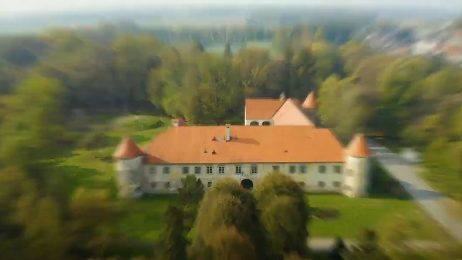 Aerial view of a historic building surrounded by trees. The structure has an orange-tiled roof with turrets at the corners, set in a lush green area. The background features a blurred forest and distant landscape.