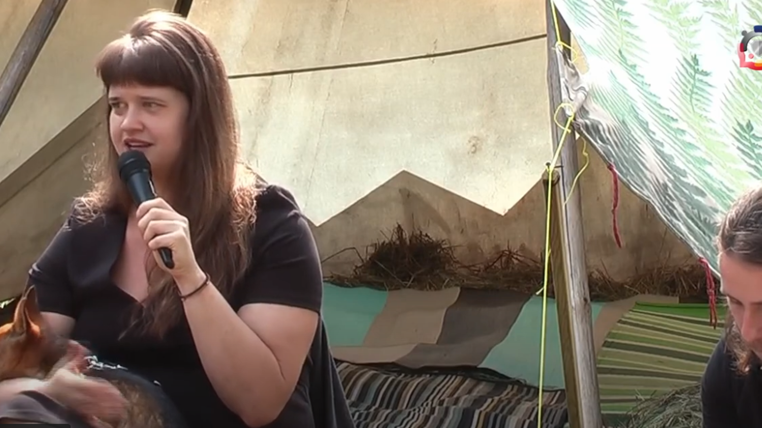 A woman with bangs holds a small dog and speaks into a microphone. She is seated in a tent or makeshift structure with patterned blankets and hay visible in the background. A man with long hair sits beside her, looking downward.