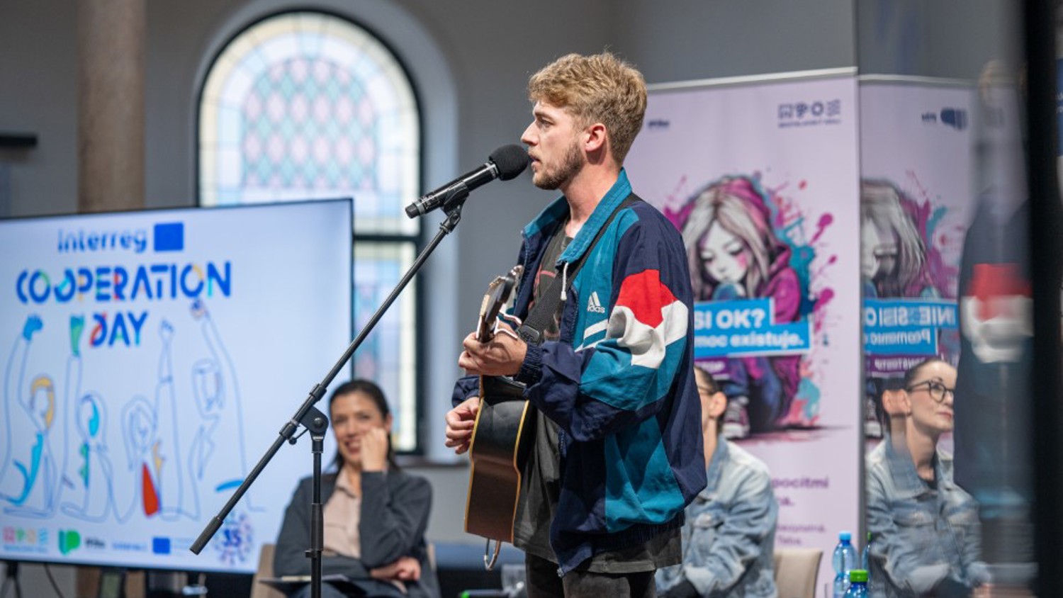 A young man in a colorful jacket sings and plays guitar on stage with a microphone. Behind him, people sit at a table, and banners and a screen reading "Cooperation Day" are visible in the background.