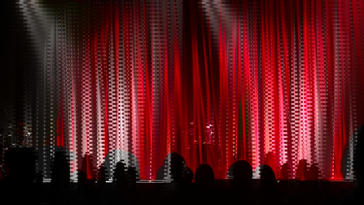 Red stage curtains are illuminated by dramatic lighting, casting bright and dark patterns. Silhouettes of audience members are visible in the foreground, and some instruments or equipment can be seen at the sides of the stage.