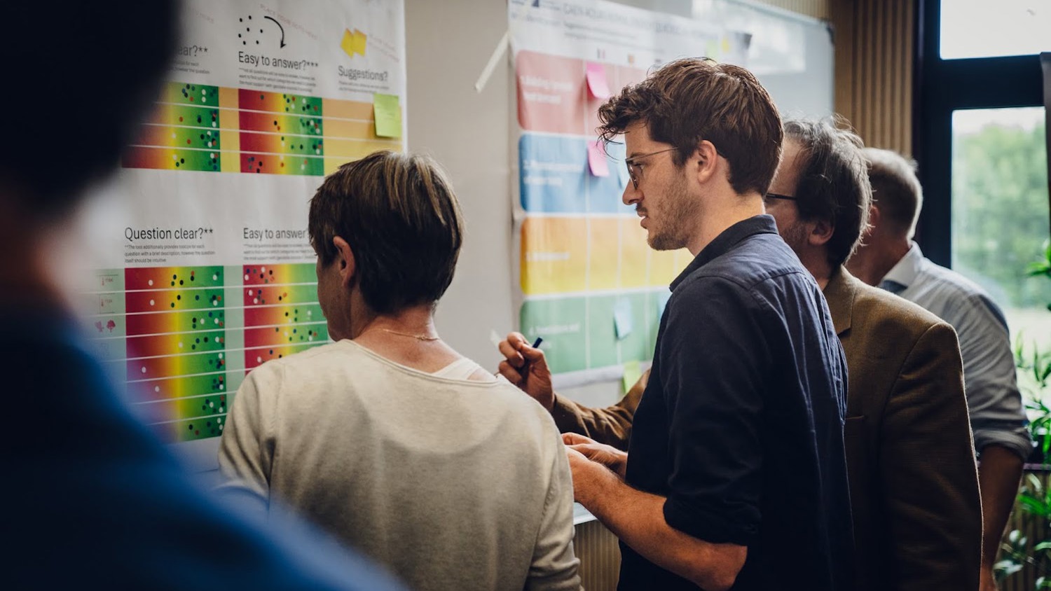 A group of people stand closely together, analyzing colorful charts and notes on a wall during a workshop or brainstorming session. One person writes on the board while others observe and discuss.