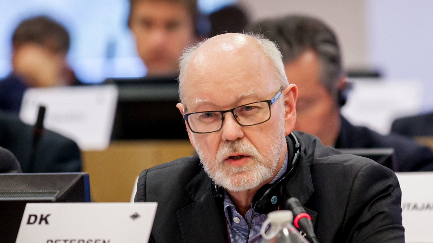 An older man with glasses and a beard speaks into a microphone at a conference table. In front of him is a nameplate labeled "DK PETERSEN" and a water bottle. Other people are seated in the background.
