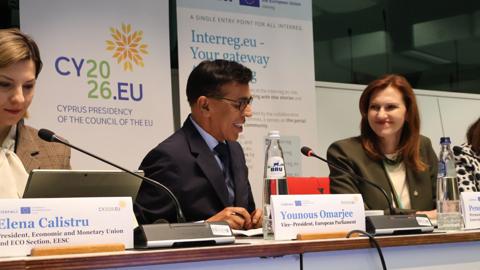 Three people sit at a conference table with microphones and nameplates. The middle person, Younous Omarjee, speaks while the others listen. Banners for Cyprus EU presidency and Interreg are visible in the background.