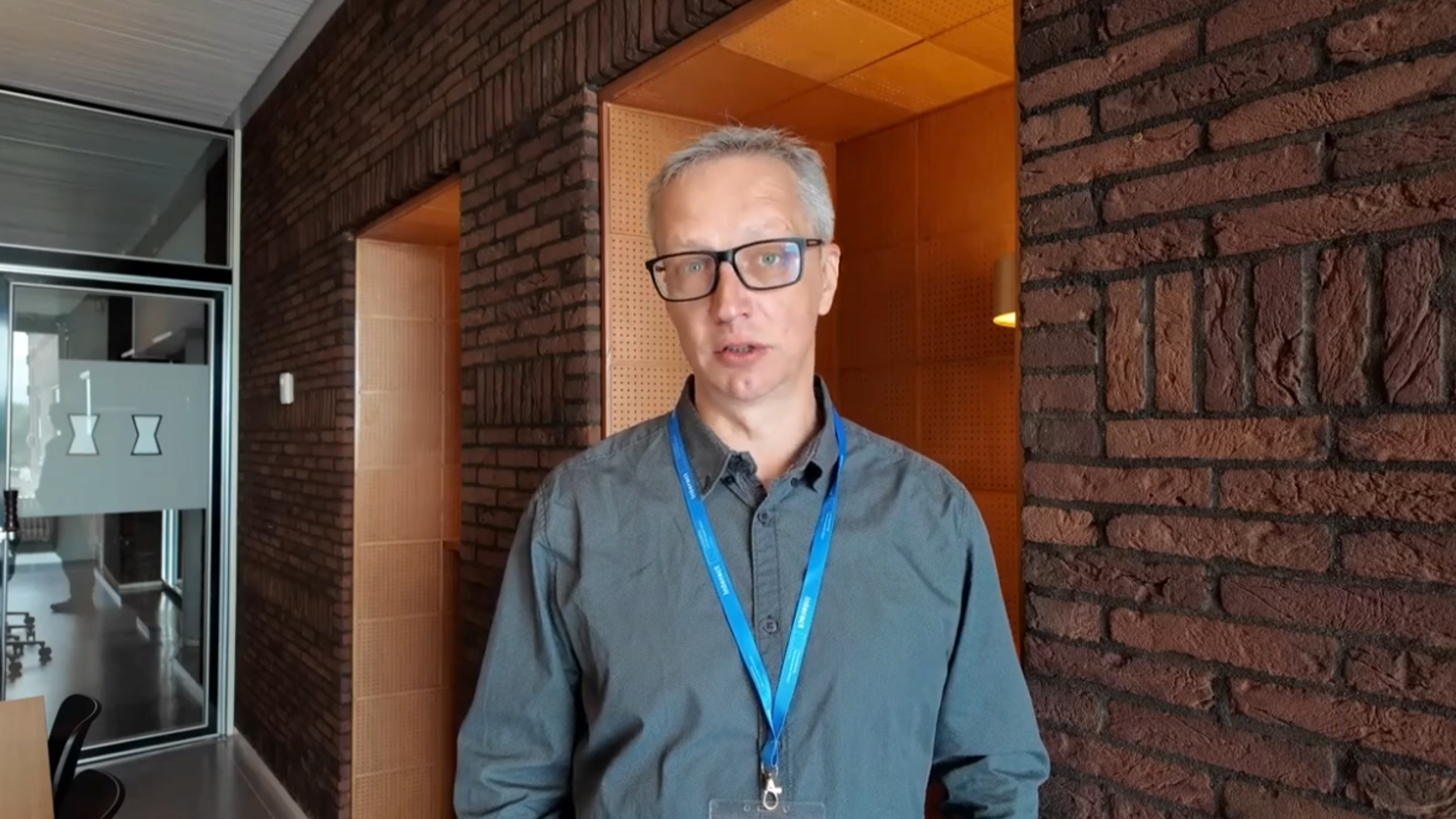 A man with short gray hair and glasses, wearing a gray shirt and blue lanyard, stands indoors against a brick wall with a hallway and glass door visible in the background.