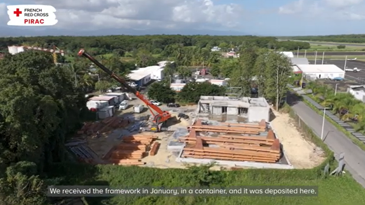 Aerial view of a construction site with a crane lifting materials, wooden beams on the ground, and trees and buildings in the background. A French Red Cross PIRAC logo is visible. A subtitle reads: “We received the framework in January...”.