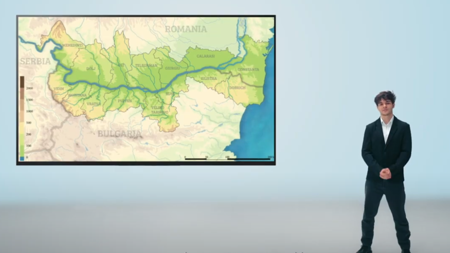 A person in a black suit stands on the right side, next to a large map showing the Danube River region, including parts of Serbia, Romania, and Bulgaria.