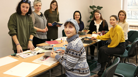 A group of women sit and stand around a table working on a crafts project with magazines, scissors, and glue in a bright, indoor room with a window and potted plant.