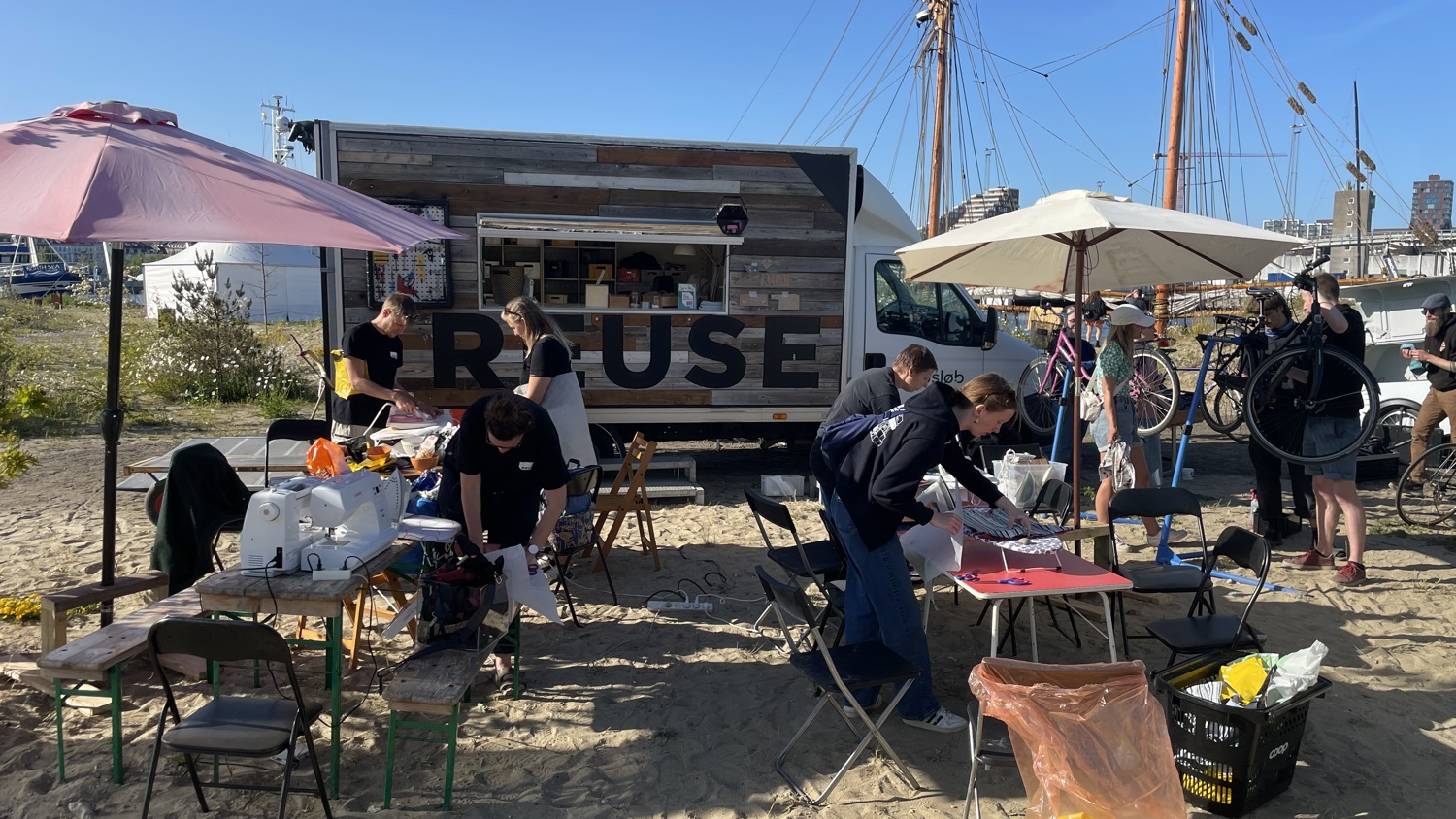 People gather around tables with supplies under umbrellas on a sandy area near a food truck, with a ship and city buildings visible in the background. Some people are seated while others stand and work on various tasks.