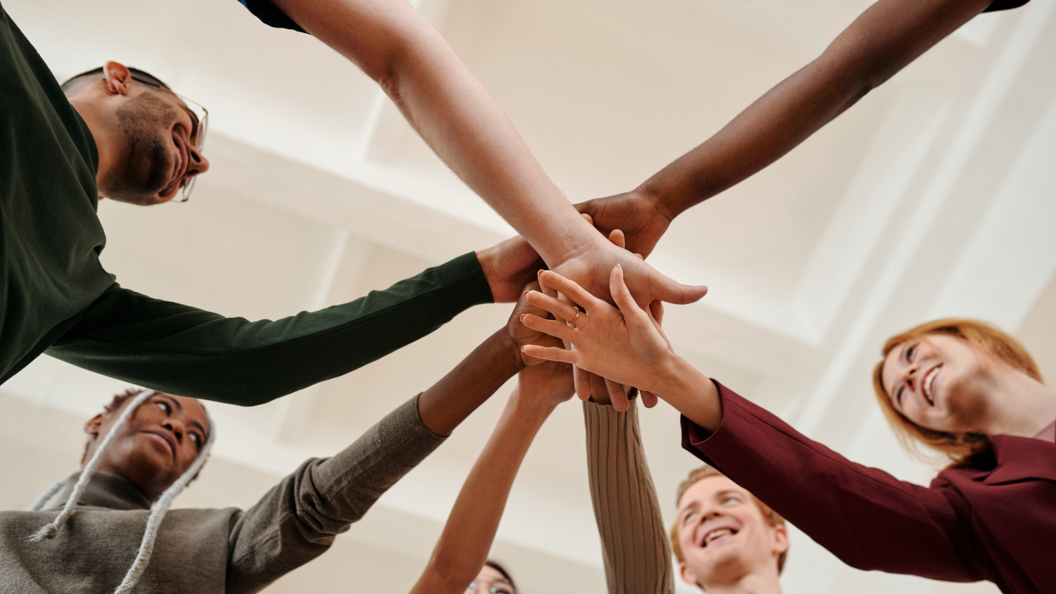 A low-angle view of a diverse group of people standing in a circle, extending their arms inward to touch hands in a gesture of unity and teamwork. They are smiling and wearing casual clothing.