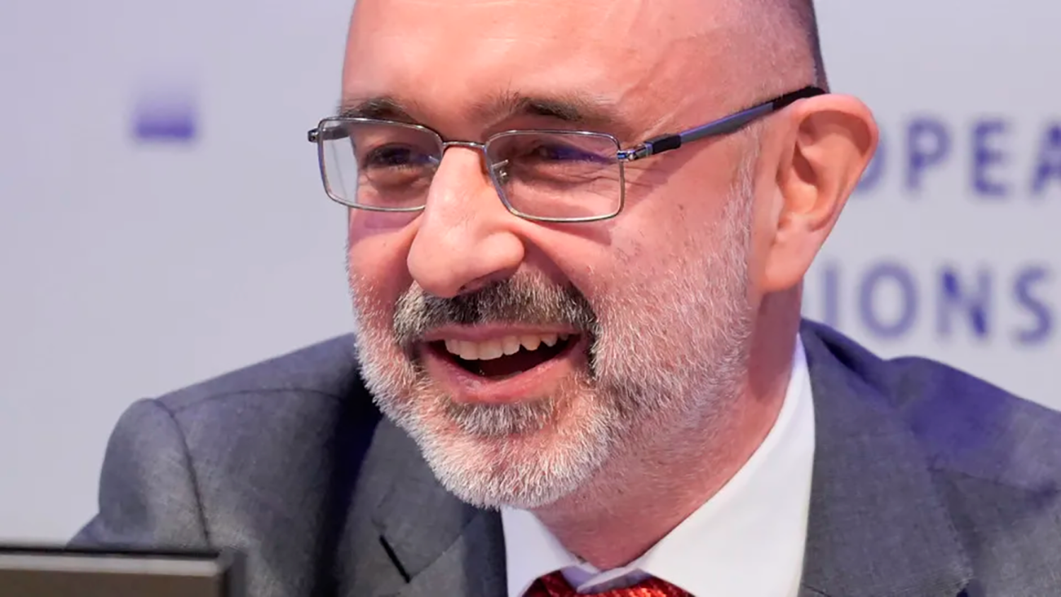 A middle-aged man with a beard and glasses smiles while wearing a suit and red tie. The background is blurred with partial, out-of-focus text.