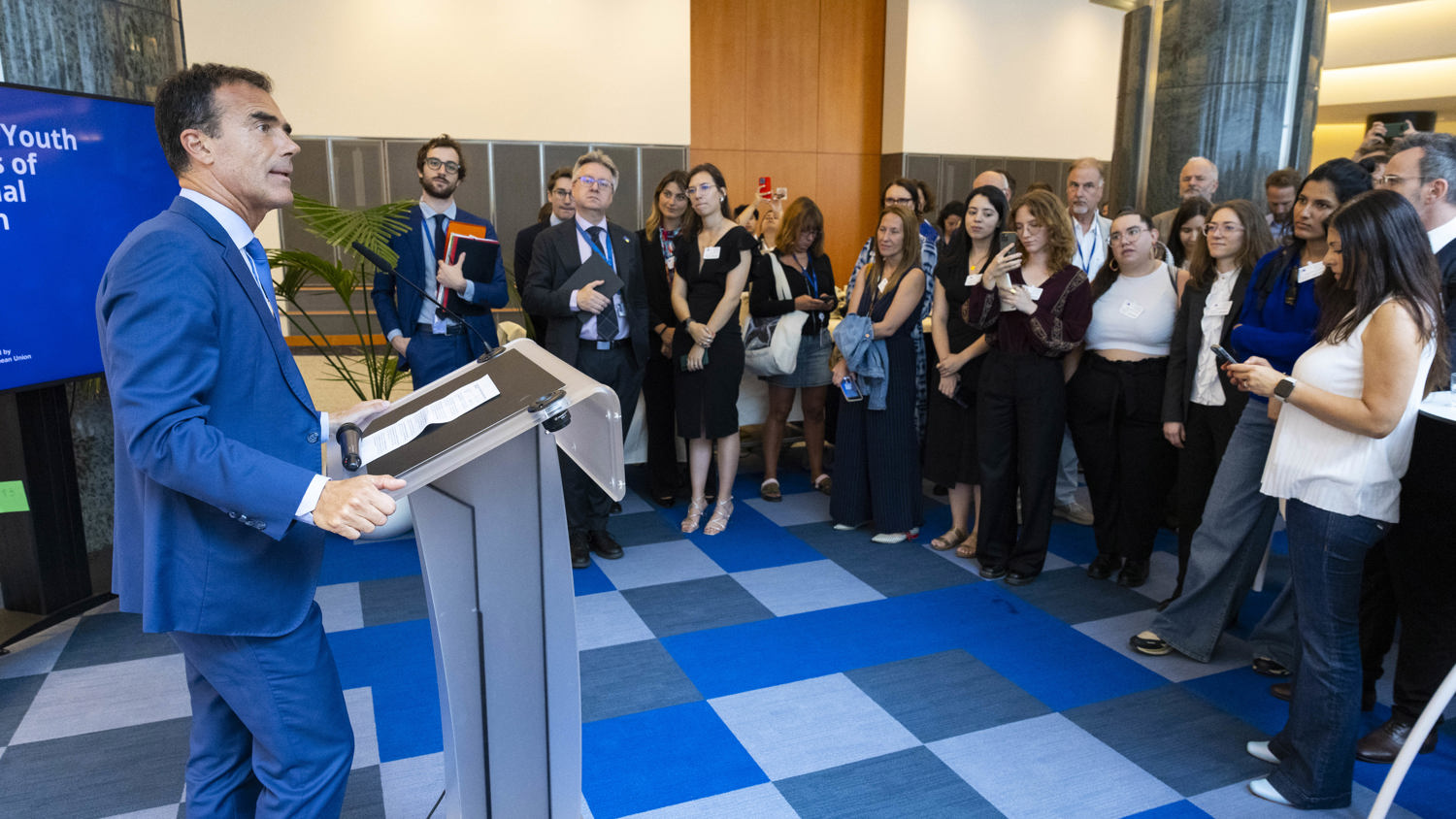 A man in a blue suit speaks at a podium to a large audience in a modern conference room. Attendees listen attentively, some standing and some holding notepads, with a presentation screen visible on the left.