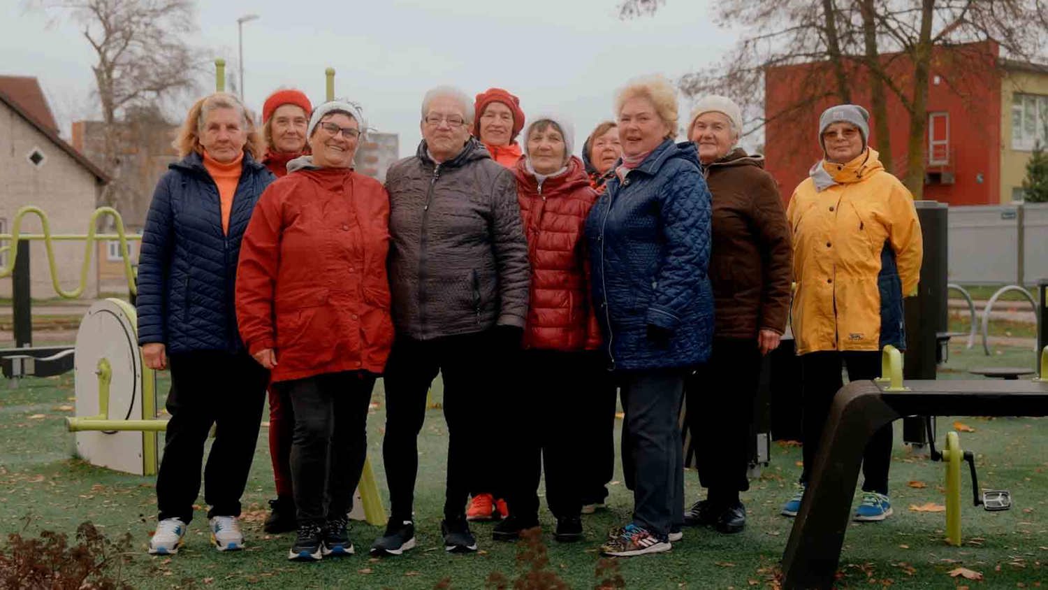 A group of older adults stands outdoors at a park gym, smiling. They are dressed warmly in coats and hats. The background features exercise equipment and trees with autumn leaves.