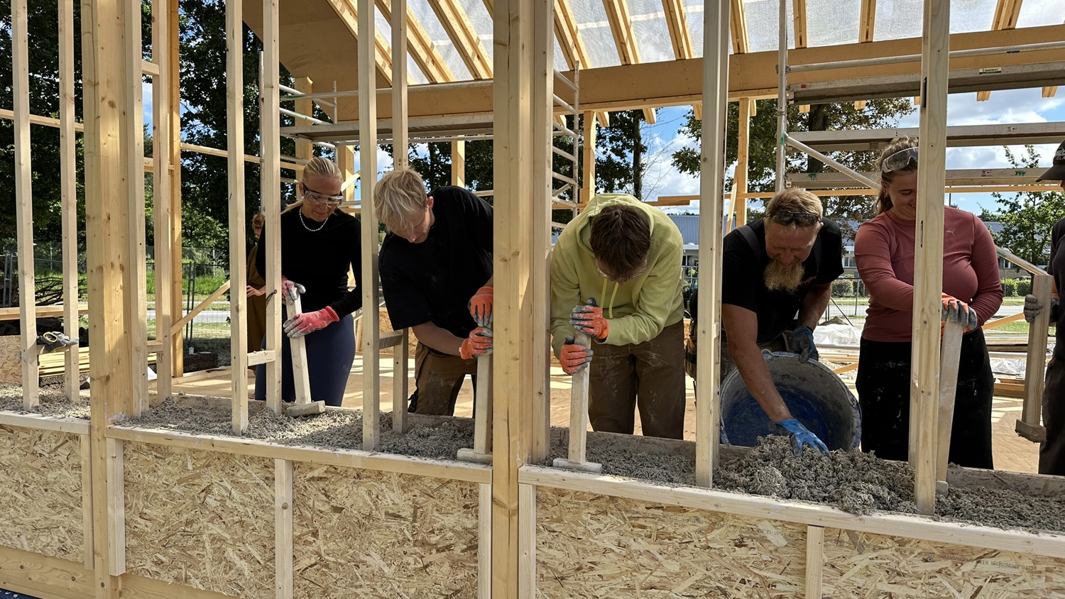 Five people work together, pouring and spreading a mixture into a wooden frame as part of building construction, with sunlight streaming through the unfinished roof structure.