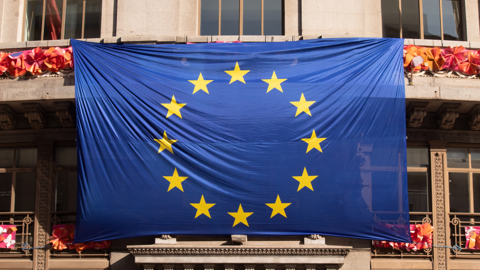 A large European Union flag with twelve yellow stars in a circle on a blue background hangs on the exterior of a building with stone walls and windows.