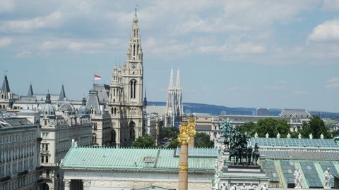 Cityscape view of Vienna with historical buildings, the tall Vienna City Hall tower, St. Stephen’s Cathedral spires in the background, and a statue of horsemen on a roof in the foreground under a partly cloudy sky.