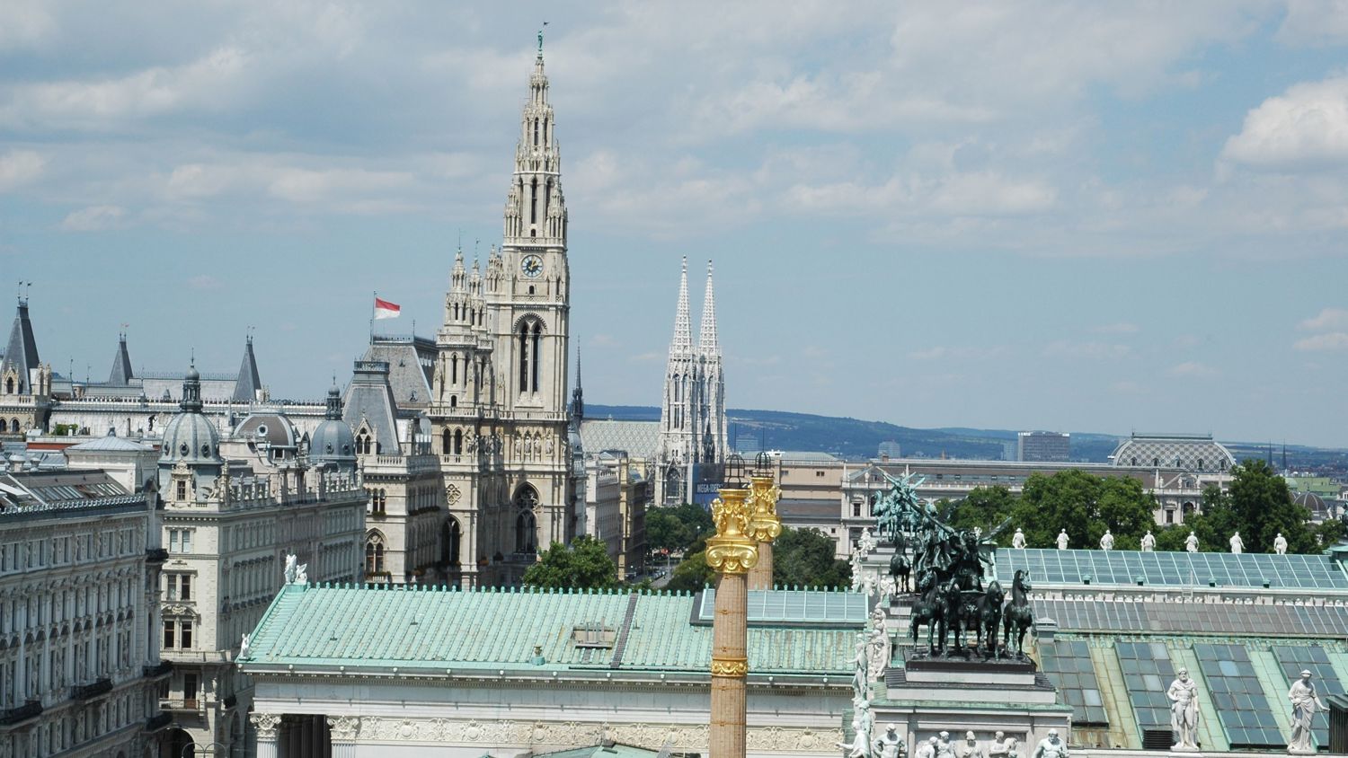 Cityscape view of Vienna with historical buildings, the tall Vienna City Hall tower, St. Stephen’s Cathedral spires in the background, and a statue of horsemen on a roof in the foreground under a partly cloudy sky.