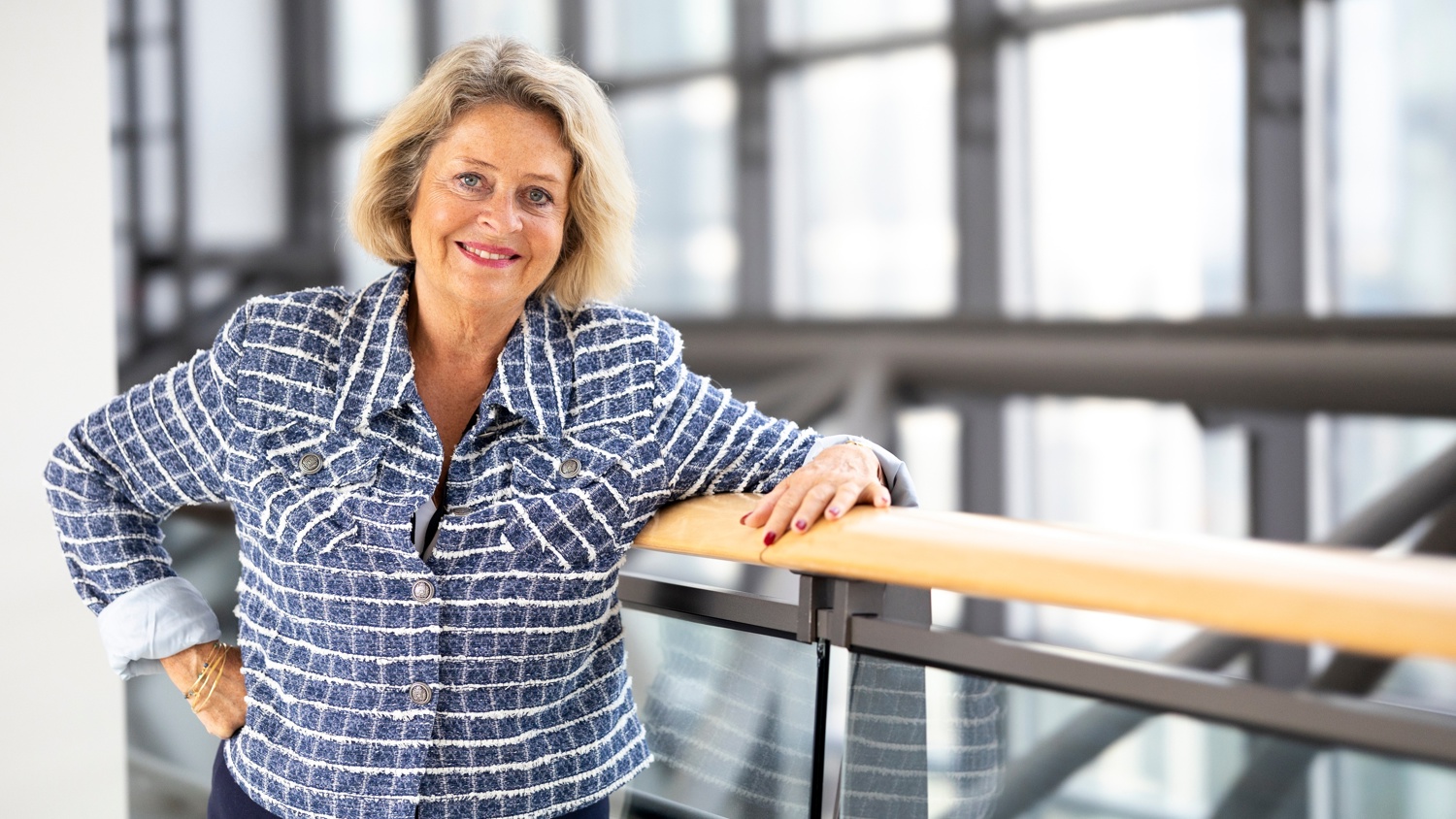 A smiling older woman with blonde hair wearing a blue and white patterned jacket stands indoors, leaning on a wooden railing with large windows in the background.