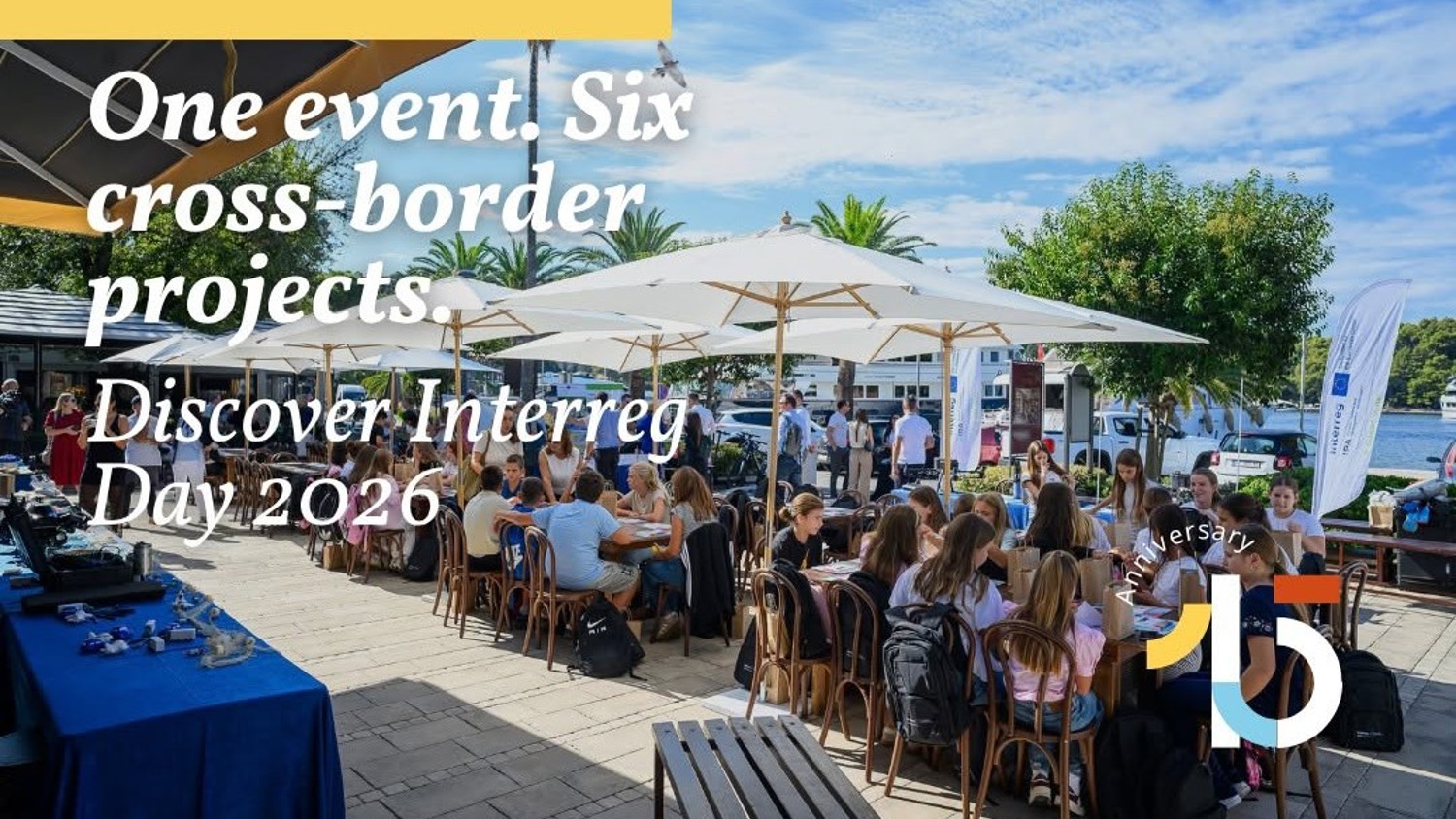 A group of people sit at outdoor tables under umbrellas near water, attending an event. Text reads, “One event. Six cross-border projects. Discover Interreg Day 2026.” Logo in the corner marks the 15th anniversary.
