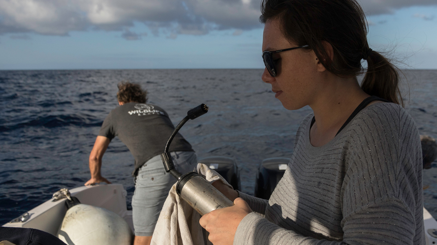 A woman in sunglasses and a gray sweater holds equipment on a boat under a cloudy sky. A man in shorts and a t-shirt stands at the boat's edge. The ocean stretches in the background.