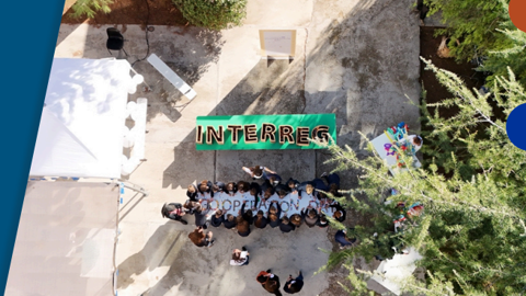 A group of people stand outdoors on a concrete path beneath a green "INTERREG" banner, surrounded by trees and tents, viewed from above.