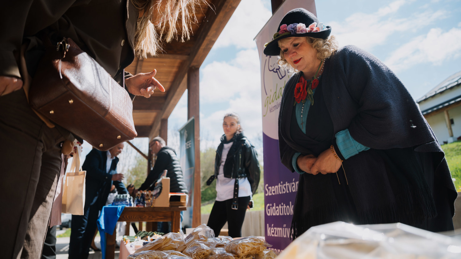 A woman in a hat and shawl stands behind a table displaying baked goods at an outdoor market. She speaks to a customer while other people browse nearby under a wooden canopy.