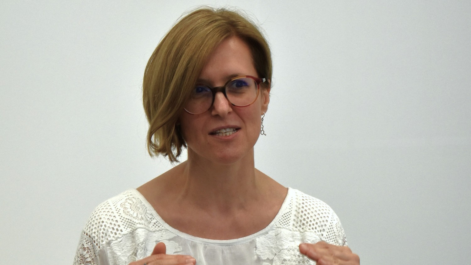 A woman with short brown hair and glasses gestures with her hands while speaking.