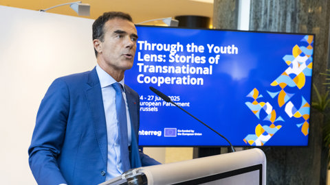 A man in a blue suit speaks at a podium in front of a screen displaying the event title "Through the Youth Lens: Stories of Transnational Cooperation" at the European Parliament in Brussels.