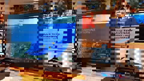 A group of people sit in front of large presentation screens in a modern conference room. The screens display text and visuals related to a climate or environmental discussion.