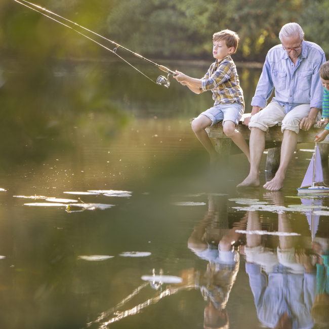 An older man sits barefoot on a wooden dock with two young boys, fishing together by a calm lake surrounded by greenery. One boy casts a fishing rod while the other looks at the water.