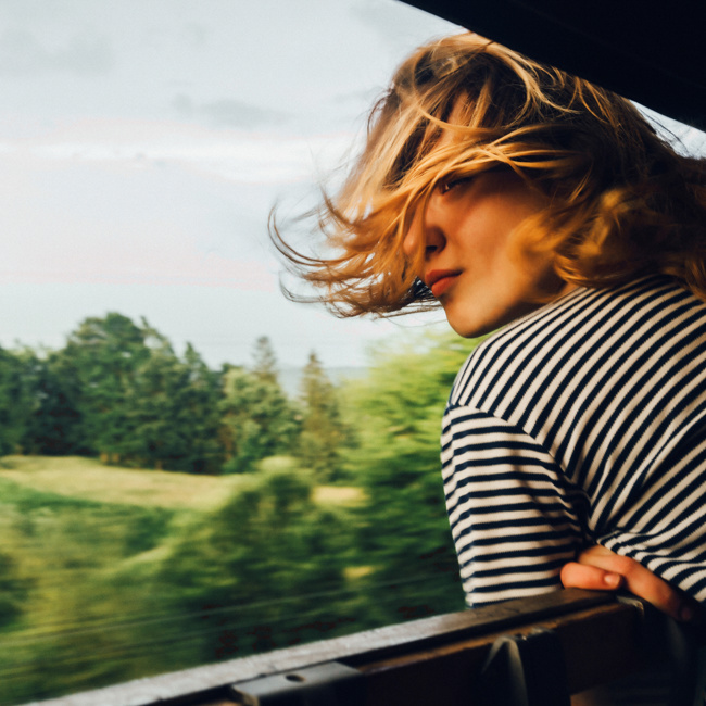 A person with shoulder-length hair, wearing a black and white striped shirt, leans out of a train window with wind in their hair, looking back, as lush green trees blur in the background.
