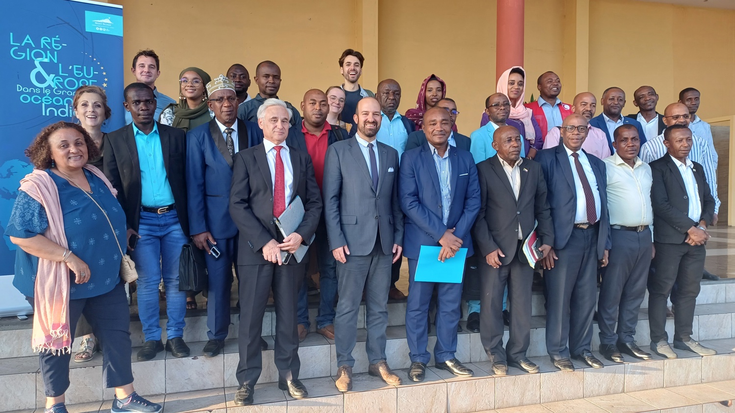 A large group of men and women in business attire stand on outdoor steps, smiling for a group photo in front of a yellow building and a blue event banner.