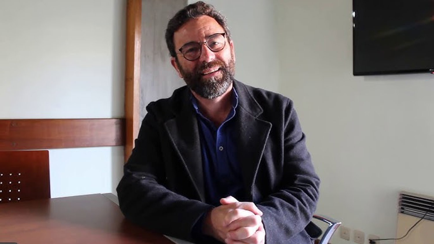 A bearded man with glasses, wearing a dark jacket and blue shirt, sits at a table smiling with his hands clasped. He is in a well-lit room with a TV on the wall and wooden furniture.