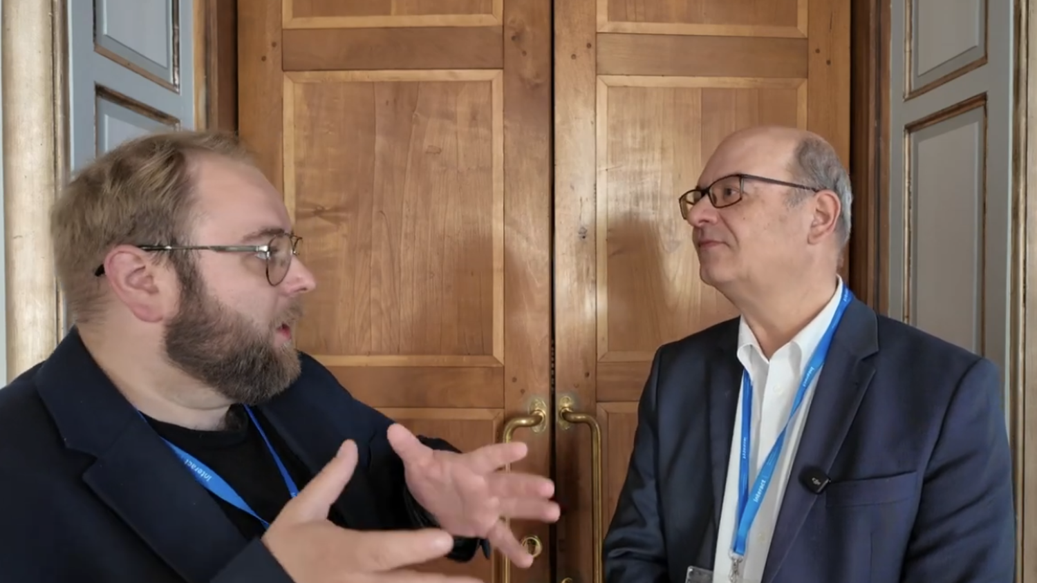 Two men in suits with blue lanyards stand talking in front of large wooden double doors; one gestures with his hands while the other listens attentively.