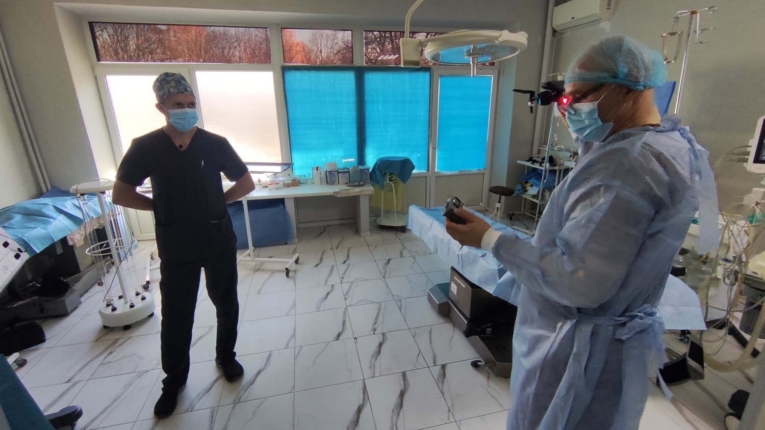 Two medical professionals in scrubs and face masks stand in a brightly lit operating room with medical equipment and blue-covered tables, one holding a device and the other standing with hands on hips.