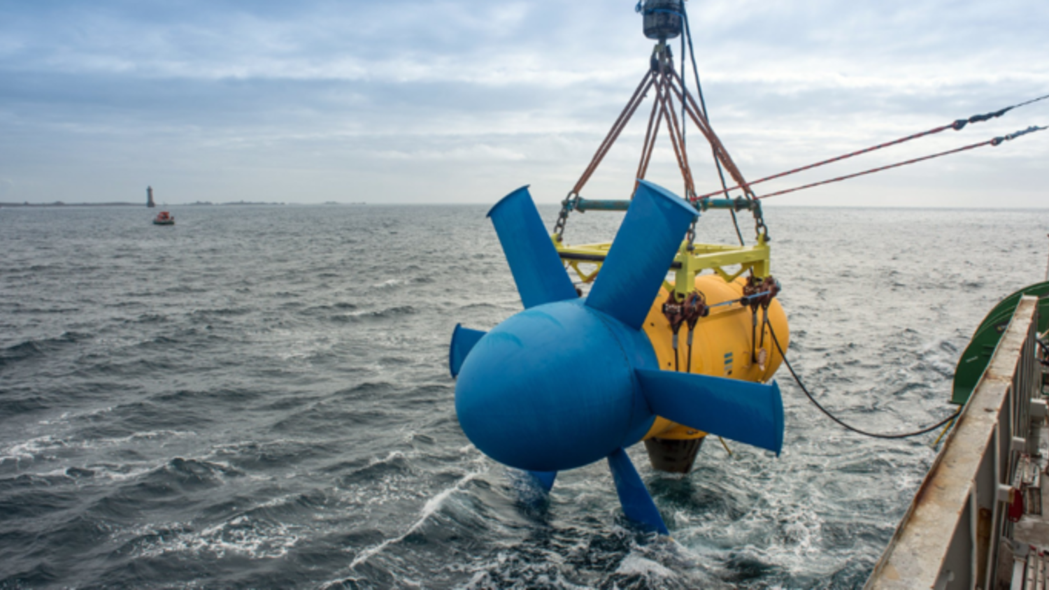A blue tidal turbine is being lowered into the ocean from a boat, supported by ropes and a crane. The sky is overcast, and there’s a distant lighthouse visible on the horizon.