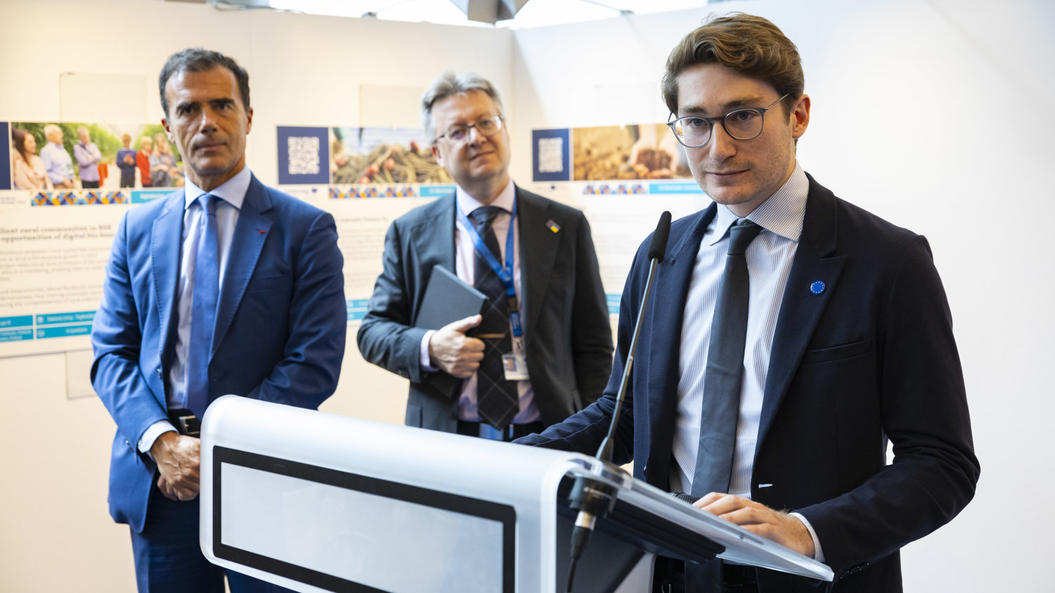 A young man in a suit speaks at a podium, while two men in suits stand behind him, listening attentively in a brightly lit room with informational posters displayed on the wall.