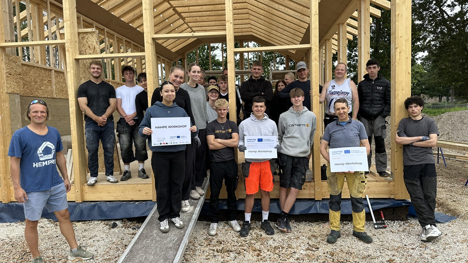 A group of people pose in front of a wooden house frame under construction. Several participants hold signs, and all appear smiling and engaged in an outdoor workshop or training event.