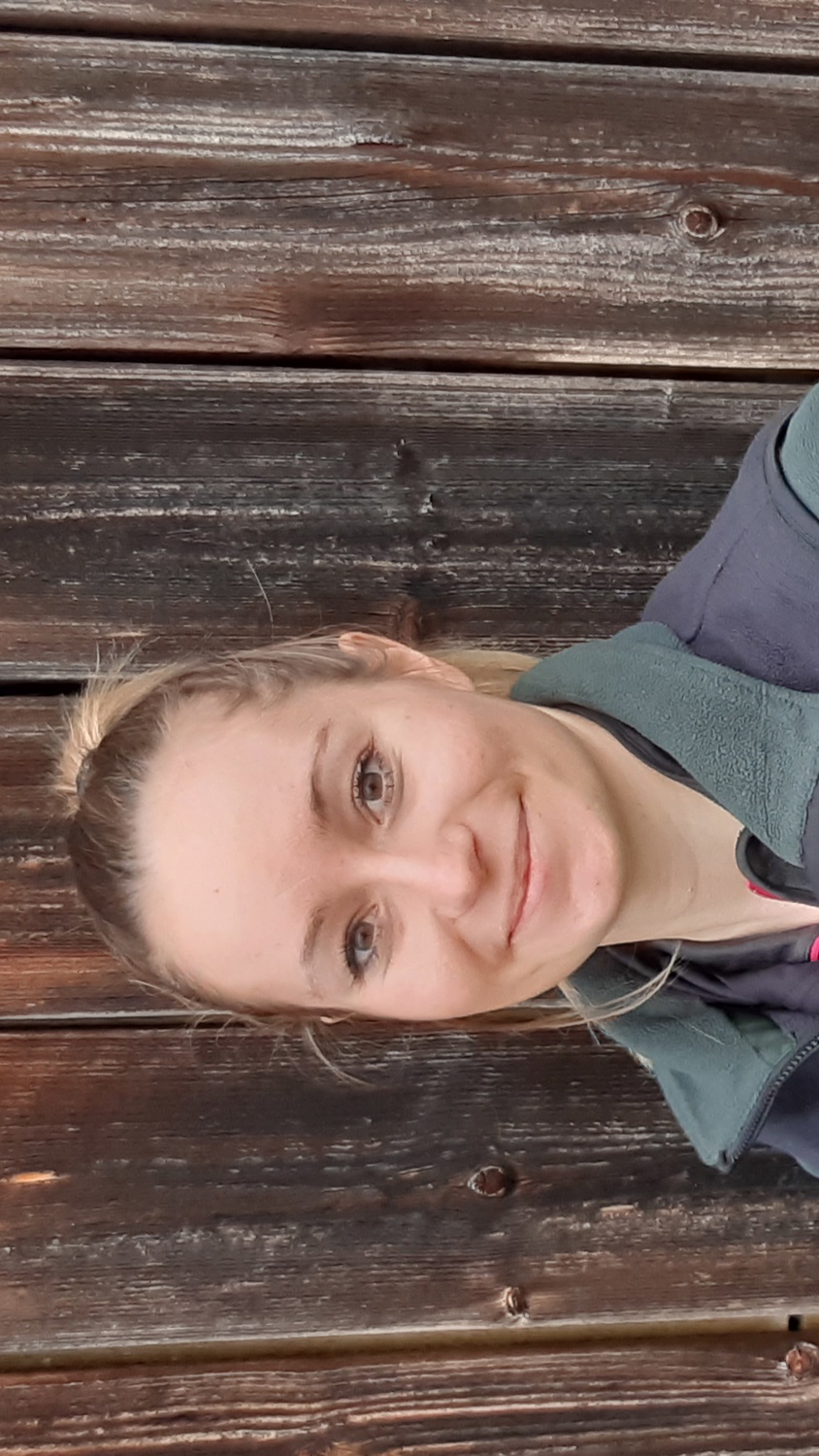 A person in a green and gray uniform jacket stands in front of a wooden wall, smiling. The jacket features a patch with blue and white checkered and black patterns, and "Forstverwaltung" text on a badge. Their hair is tied back.