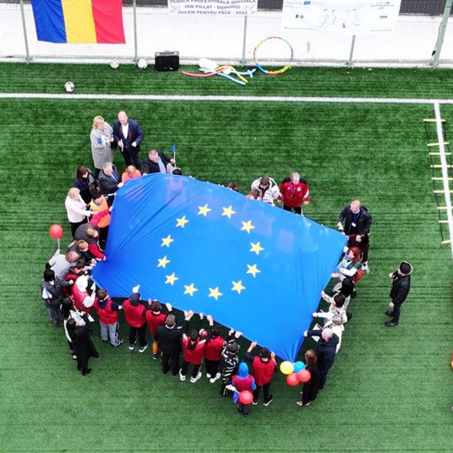 A group of people, including children holding colorful balloons, are gathered on a green field holding a large European Union flag. A Romanian flag and hoops are in the background on a fence.