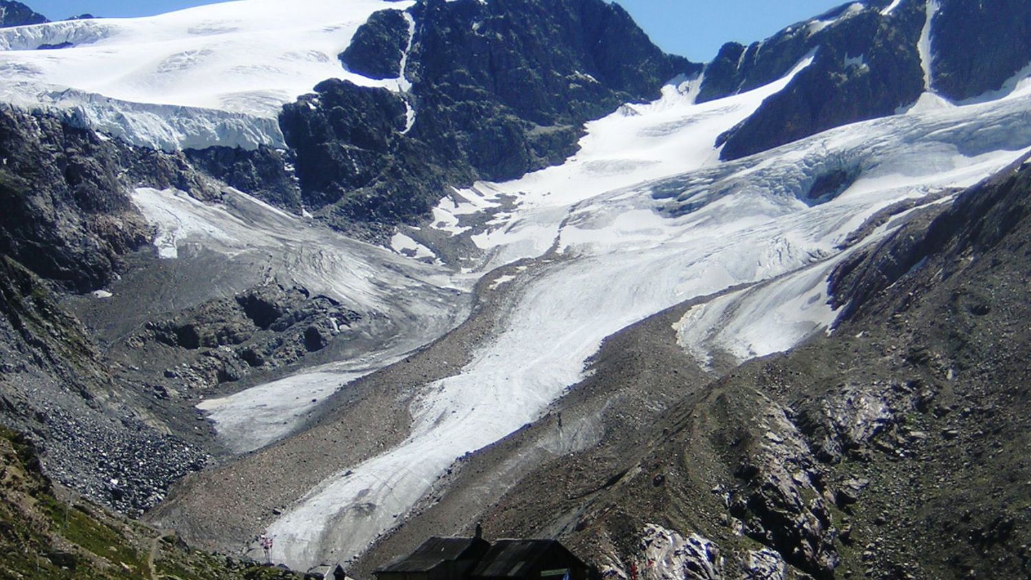 A rugged mountain landscape with a glacier flowing between rocky peaks under a clear blue sky. In the foreground, small wooden huts rest on a grassy slope.