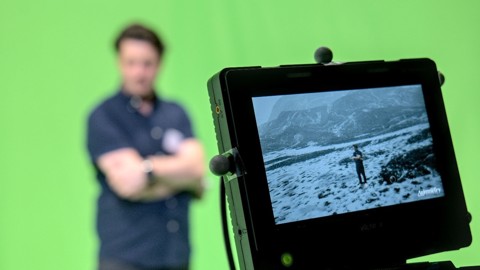 A camera monitor displays a snowy landscape with a person standing in it, while a man stands blurred in front of a green screen in the background, suggesting visual effects filming.
