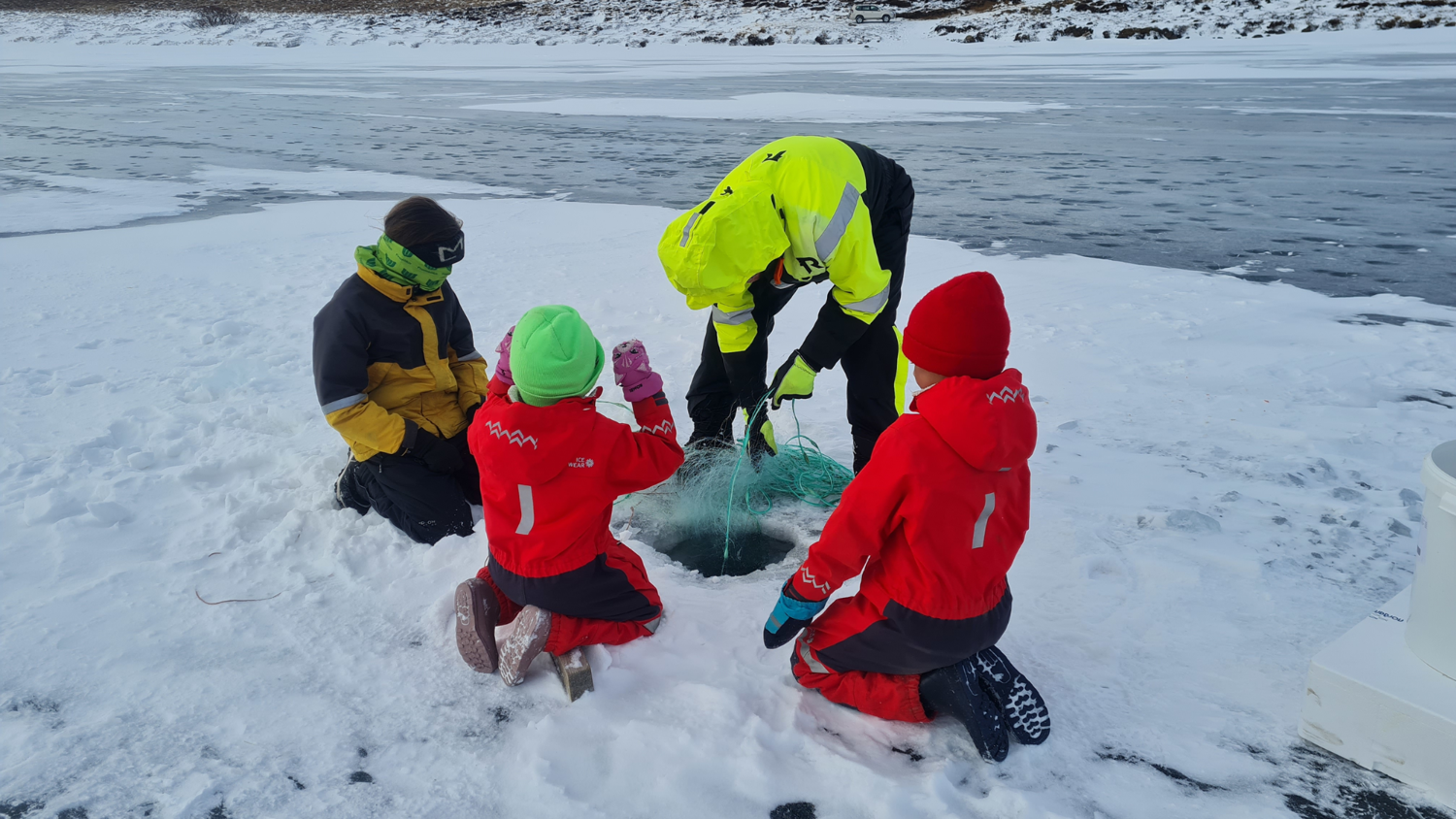 People in winter clothing gather around a hole in the ice, engaged in ice fishing. Behind them is a frozen lake. Snow covers the ground and the landscape is wintry. The group includes adults and children wearing bright jackets.