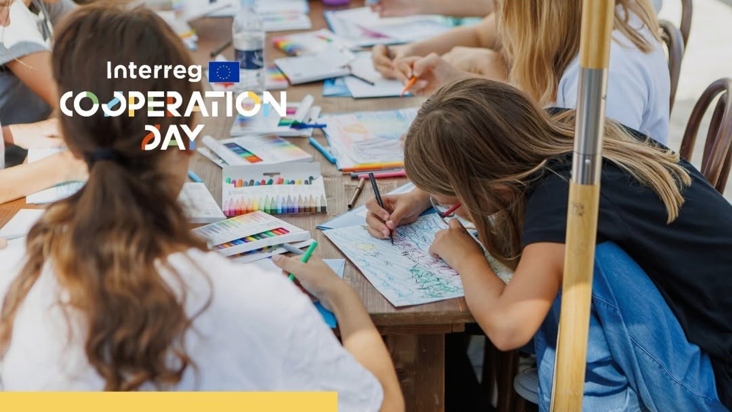Children and adults sit around a table outdoors, coloring and drawing with markers and colored pencils. The words “Interreg Cooperation Day” are overlaid on the image in white text.
