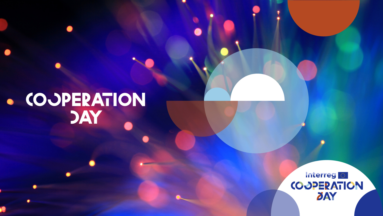 A young woman sits at a desk with a laptop, smiling and talking to another person who is partially visible, suggesting a friendly conversation or a collaborative work setting.