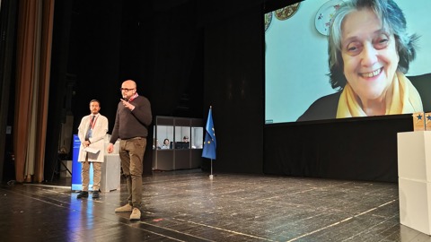 Two people stand on a stage, one speaking into a microphone, while a large screen shows a smiling woman on a video call. A blue flag and two people in a booth are visible in the background.